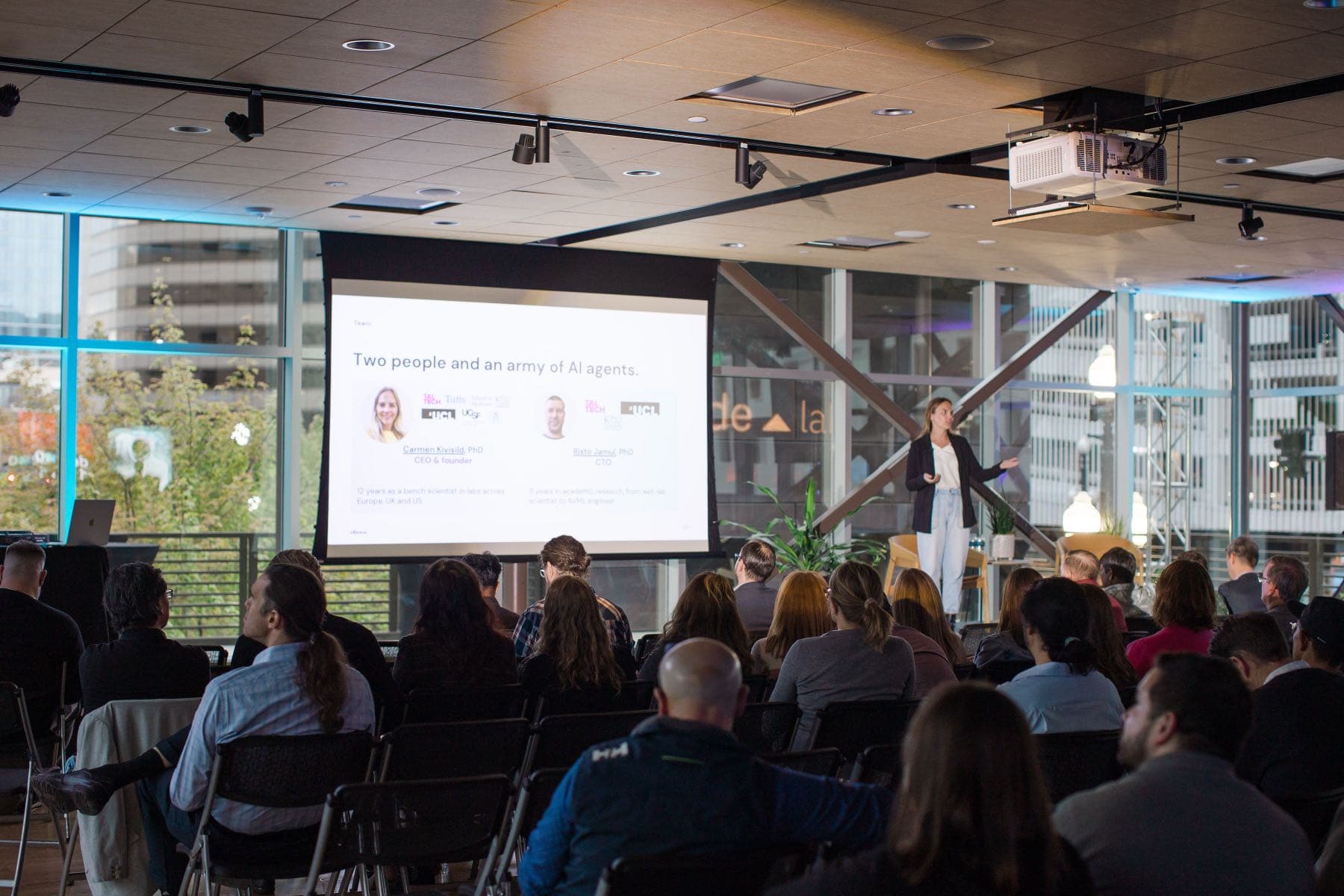 Carmen Kivisild presenting Elnora's pitch at Altitude Lab Demo Day, a slide reading “Two people and an army of AI agents” on the screen beside her, with a seated audience filling the room.
