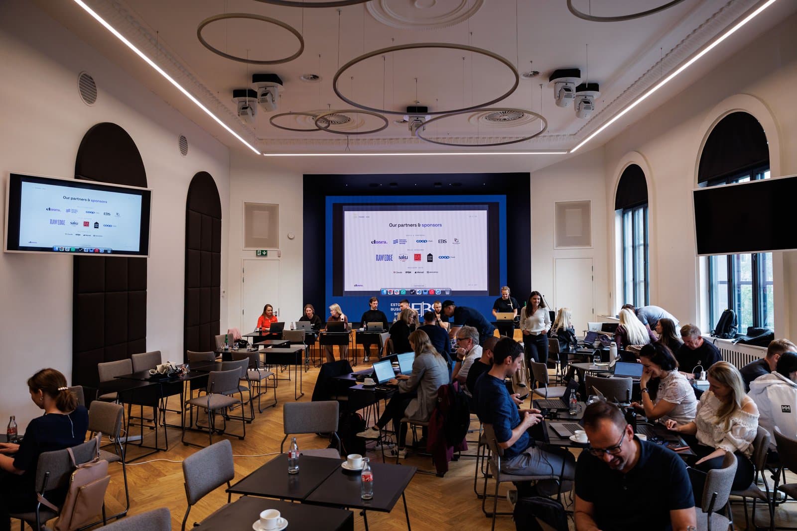 Build Day at Estonian Business School: teams working at laptops across the hall, with the partners and sponsors slide on the main screen.