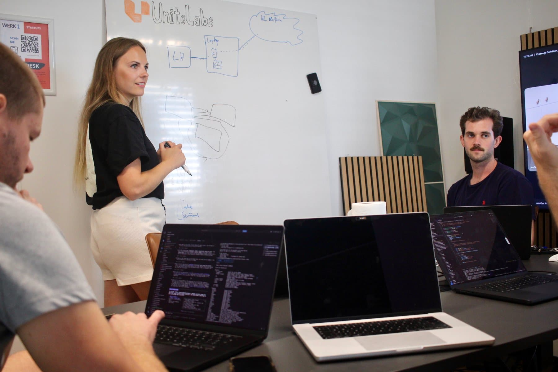 Carmen Kivisild at the whiteboard in the UniteLabs office in Munich, sketching the integration while the team works at their laptops.