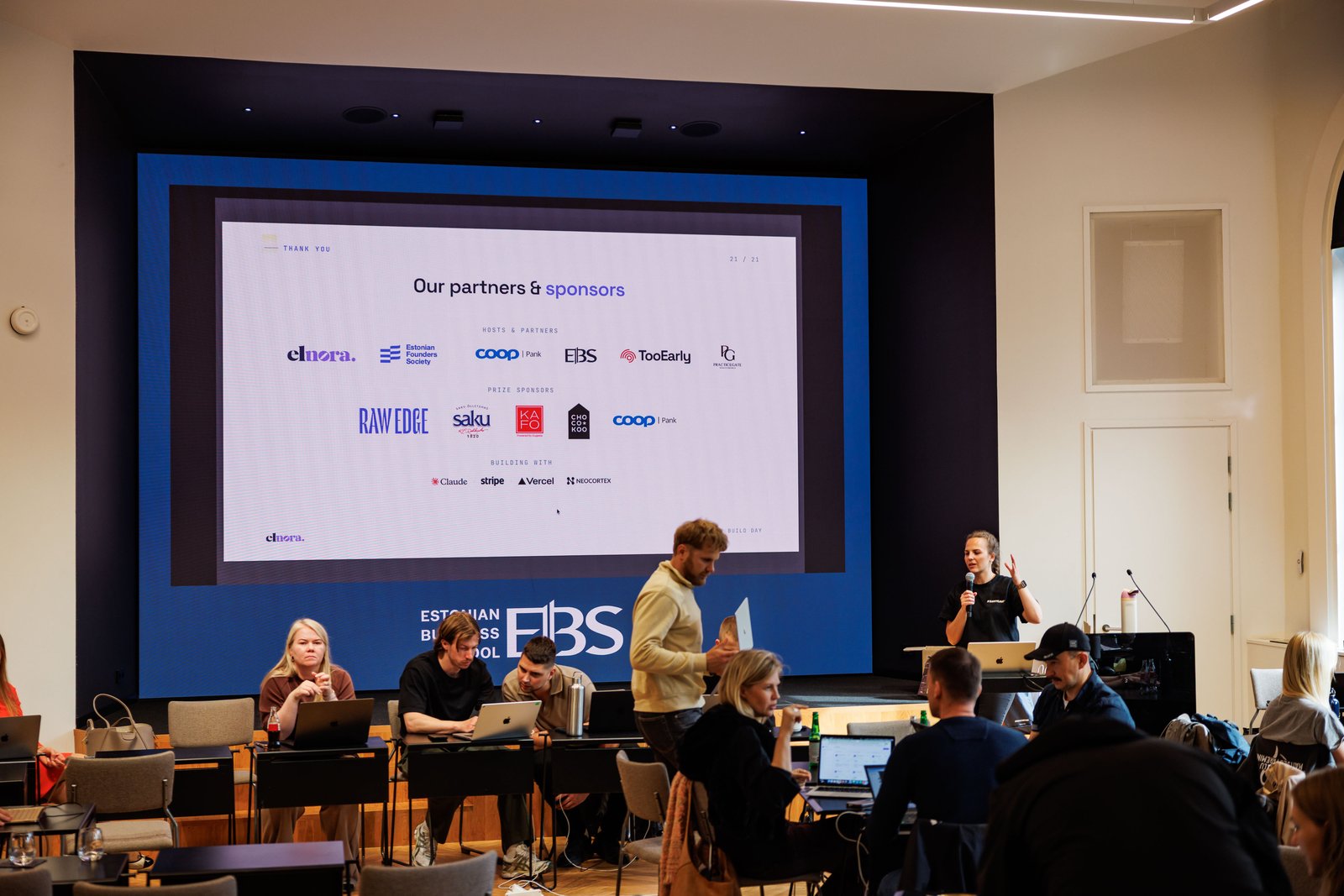 A host speaking from the stage at Build Day while teams work at laptops in front.
